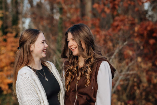 Sisters laughing together, smiling at one another, fall foliage backdrop